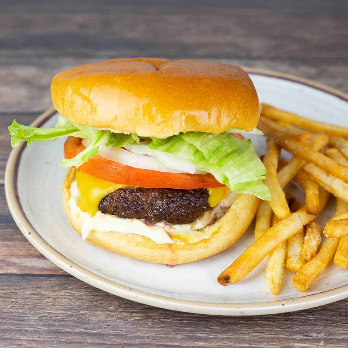 Air fryer cheeseburger on a brioche bun topped with lettuce, tomato, and melted American cheese served with a side of golden french fries on a white plate.
