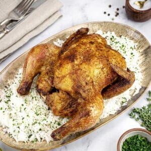 Overhead view of a roasted air fryer cornish game hen on a gold oval tray with white rice, fresh thyme, peppercorns, and seasonings nearby.