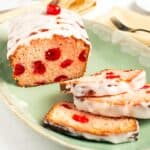 Square close up of Cherry Bread showing maraschino cherries inside the pink crumb, drizzled with white glaze and topped with a cherry, three slices fanned on a green platter.