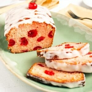 Square close up of Cherry Bread showing maraschino cherries inside the pink crumb, drizzled with white glaze and topped with a cherry, three slices fanned on a green platter.