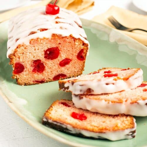 Square close up of Cherry Bread showing maraschino cherries inside the pink crumb, drizzled with white glaze and topped with a cherry, three slices fanned on a green platter.