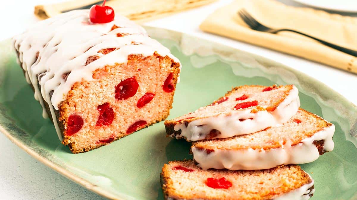 Wide view of a Cherry Bread loaf and slices on a green platter, white glaze drizzled across the top with maraschino cherries inside the pink crumb.
