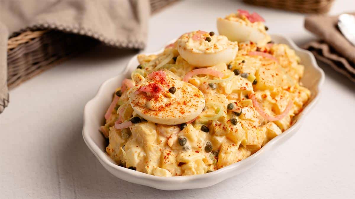 Creamy Deviled Egg Potato Salad in a white oval serving dish, garnished with deviled egg halves, pickled red onions, capers and paprika, on a light surface with a linen napkin.