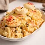 Close up of creamy Deviled Egg Potato Salad in a white dish, topped with deviled egg halves, pickled red onions, capers and paprika.