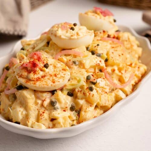 Close up of creamy Deviled Egg Potato Salad in a white dish, topped with deviled egg halves, pickled red onions, capers and paprika.