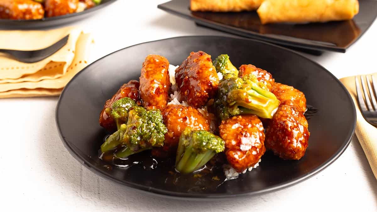 Plated General Tso's chicken and broccoli in a glossy sauce over white rice in a black bowl, with egg rolls in the background.