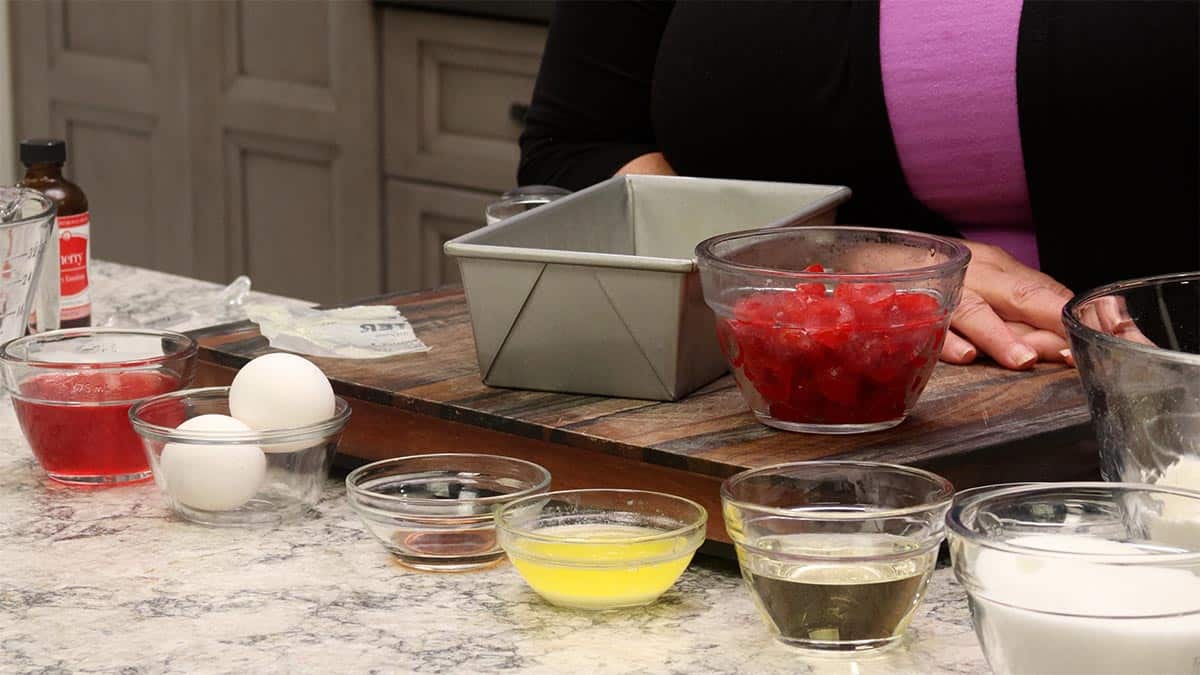 Ingredients for Cherry Bread arranged on a marble counter, including a loaf pan, jar of maraschino cherries, flour, sugar, eggs, butter, oil, half and half, and small bowls of leavening, salt, vanilla, and cherry baking emulsion.