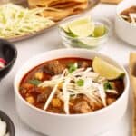 Close up of Pork Pozole Rojo in a white bowl topped with shredded cabbage, onion, cilantro, and a lime wedge, with lime wedges and crispy tortillas in the background.