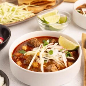 Close up of Pork Pozole Rojo in a white bowl topped with shredded cabbage, onion, cilantro, and a lime wedge, with lime wedges and crispy tortillas in the background.