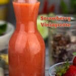Glass carafe of bright pink strawberry vinaigrette dressing on a white wooden surface, with a side salad in a white bowl, a mason jar of granola, fresh strawberries in a small bowl, and a green plaid napkin in the background.