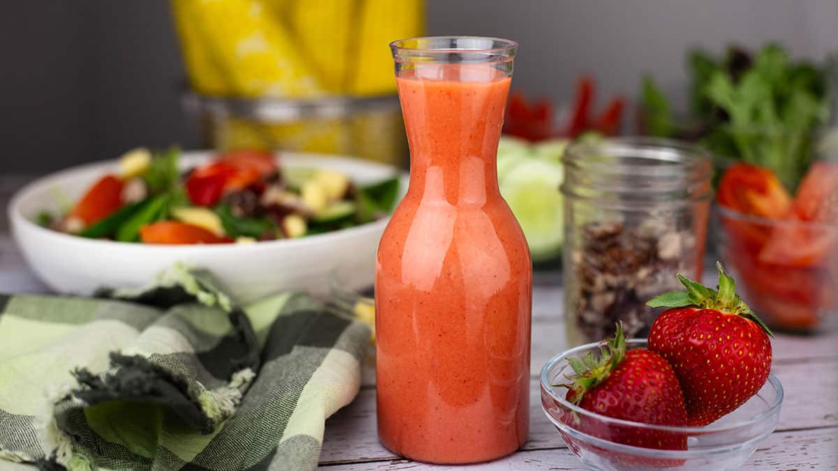 Tall glass carafe filled with creamy strawberry vinaigrette dressing on a rustic white table, beside a salad bowl, a jar of granola, ripe strawberries, and a yellow blender pitcher in the background.