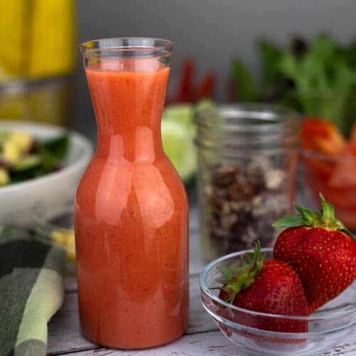 Square close up of a glass carafe of strawberry vinaigrette dressing on a white wooden surface with fresh strawberries in a small clear bowl and a mason jar of granola alongside.