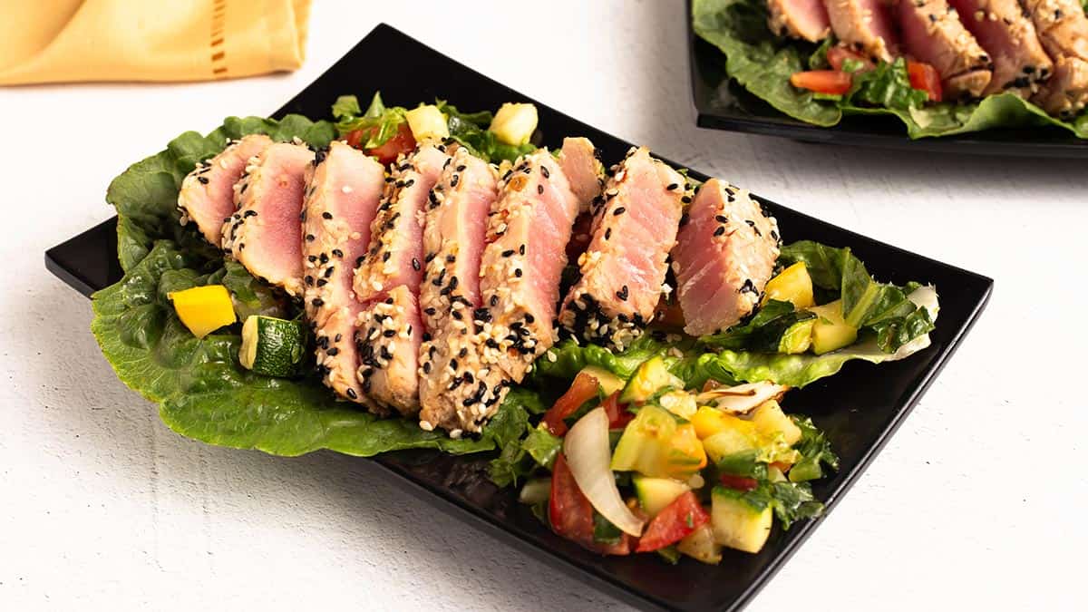 Sliced sesame crusted Air Fryer Tuna Steaks fanned over green lettuce with a chopped squash, tomato, and onion salad on a black plate.