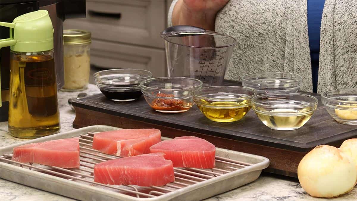 Ingredients for Air Fryer Tuna Steaks, raw tuna steaks on a rack with bowls of oil, soy sauce, and seasonings, an oil sprayer, and an onion.
