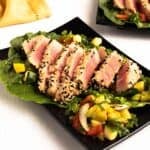 Sliced sesame crusted Air Fryer Tuna Steaks on lettuce with a colorful chopped squash and tomato salad on a black plate.