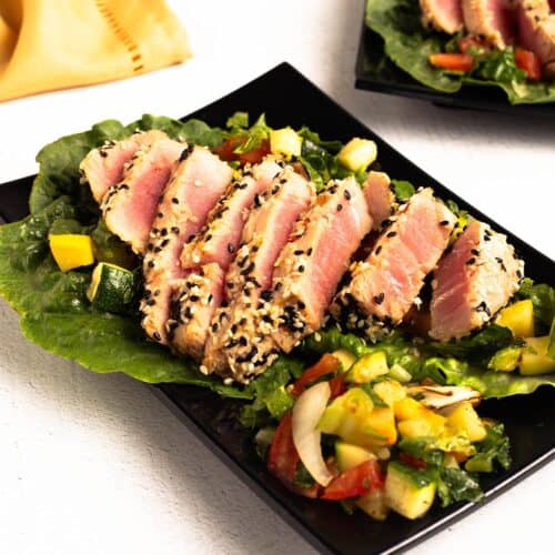 Sliced sesame crusted Air Fryer Tuna Steaks on lettuce with a colorful chopped squash and tomato salad on a black plate.