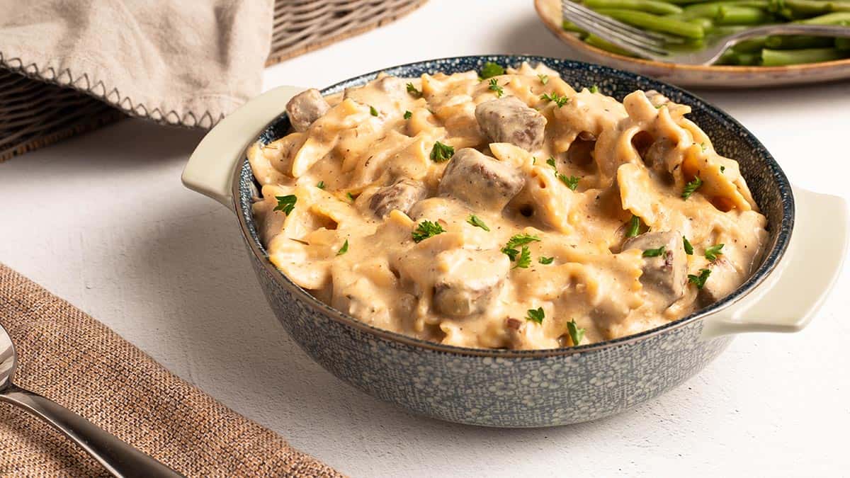 Square photo of creamy beef tips and pasta in a blue speckled bowl, topped with fresh parsley, with green beans in a dish behind it.