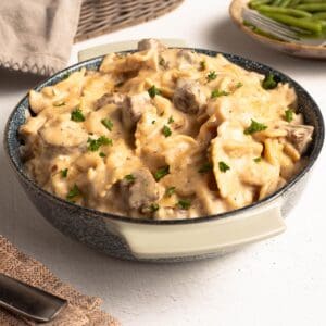 Square photo of creamy beef tips and pasta in a blue speckled bowl, topped with fresh parsley, with green beans in a dish behind it.