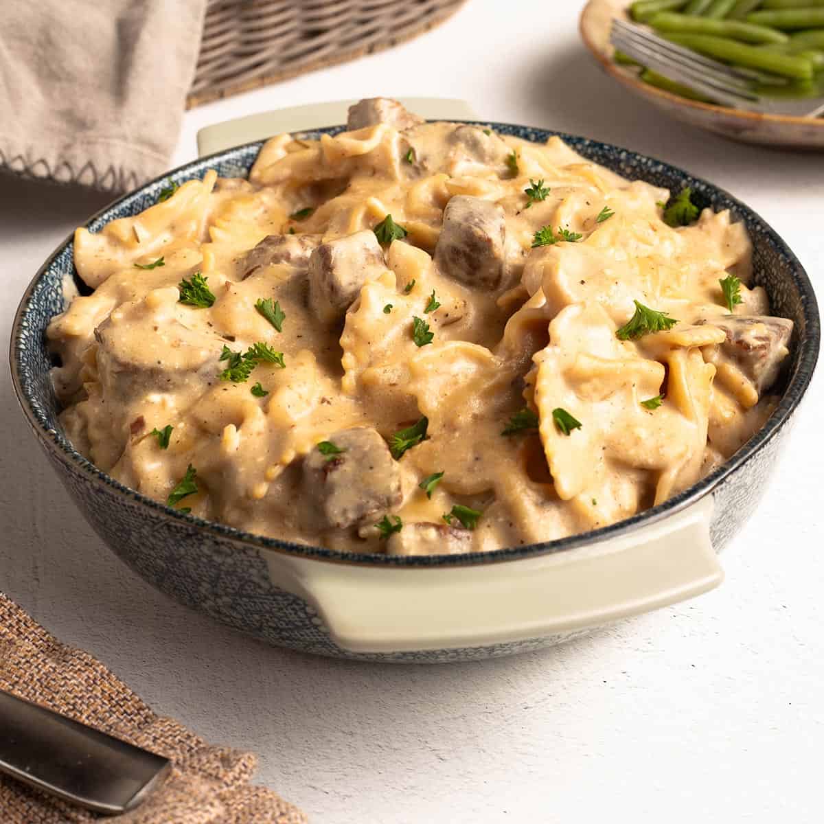Square photo of creamy beef tips and pasta in a blue speckled bowl, topped with fresh parsley, with green beans in a dish behind it.