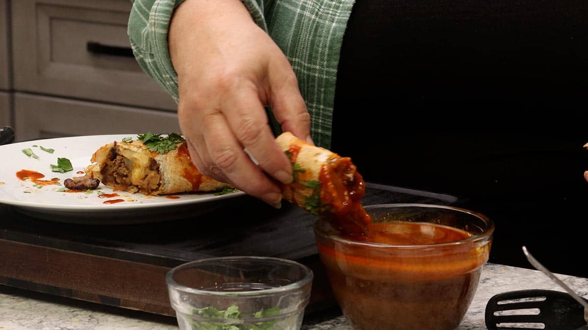 A hand dipping a crispy One-Pot Burrito into a glass bowl of au jus, with a second burrito and cilantro on a white plate.
