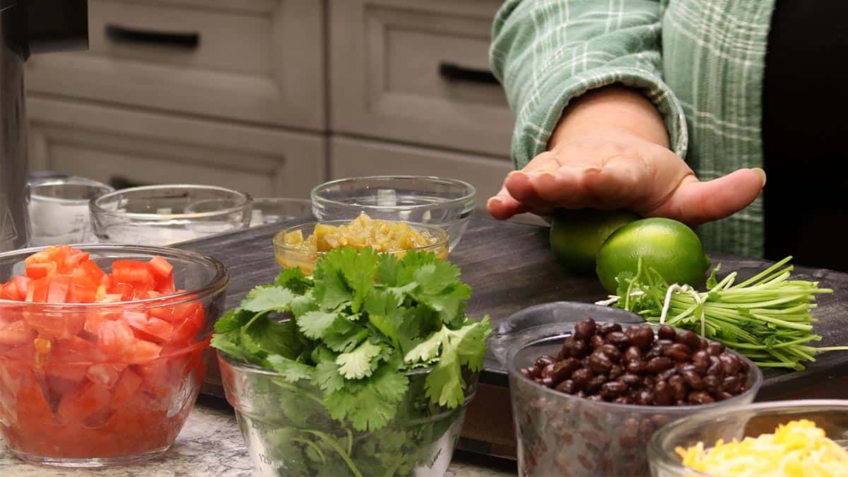 Glass bowls of chopped tomatoes, cilantro, green chiles, black beans, and shredded cheese with two limes on a wood board, the ingredients for One-Pot Burritos.