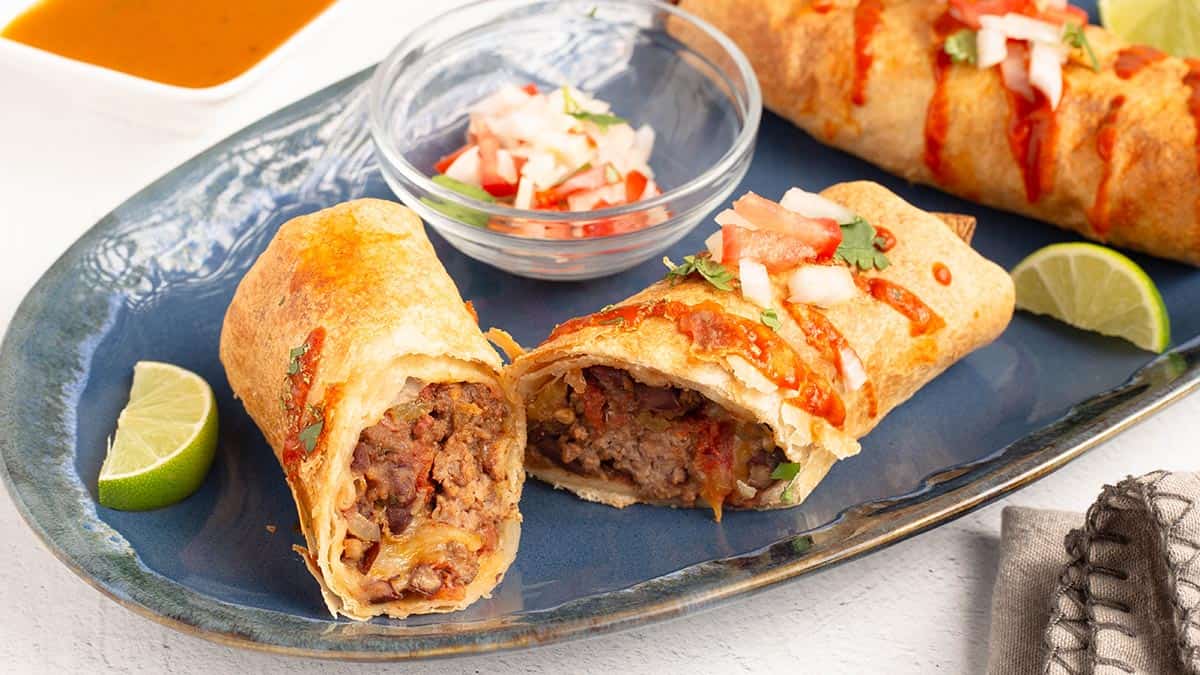 Two crispy One-Pot Burritos on a blue plate, one cut to reveal the beef and bean filling, served with lime wedges, pico de gallo, and au jus.