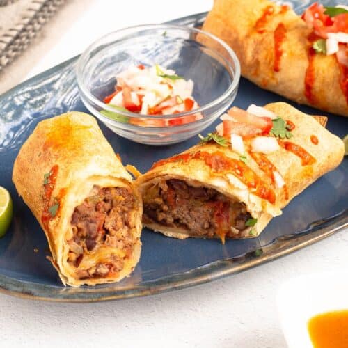 Two golden One-Pot Burritos on a blue plate topped with hot sauce and pico de gallo, one cut open to show the cheesy beef and bean filling.
