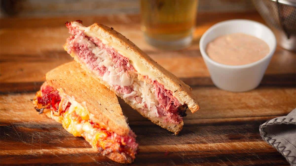 A halved Reuben sandwich stacked on a wooden board showing melted Swiss cheese, corned beef, and sauerkraut on toasted rye, with a bowl of Thousand Island dressing alongside.