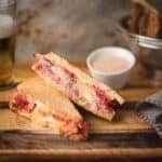 A Reuben sandwich on toasted rye cut in half and stacked on a wooden board, served with a small bowl of Thousand Island dressing and a glass of beer.