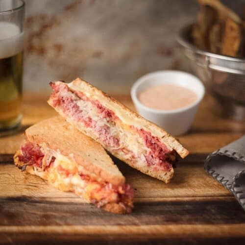 A Reuben sandwich on toasted rye cut in half and stacked on a wooden board, served with a small bowl of Thousand Island dressing and a glass of beer.
