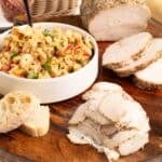 Low roasted turkey breast on a wooden serving board with thick peppered slices, thin shaved slices, a bowl of creamy turkey salad garnished with parsley, and toasted baguette rounds.