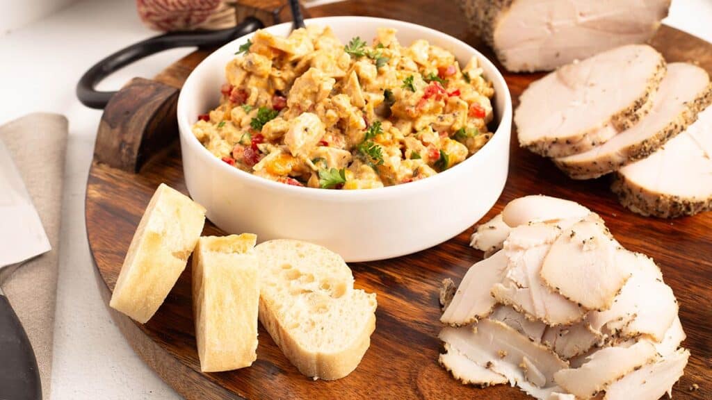 Low roasted turkey breast on a wooden serving board with thick peppered slices, thin shaved slices, a bowl of creamy turkey salad garnished with parsley, and toasted baguette rounds.