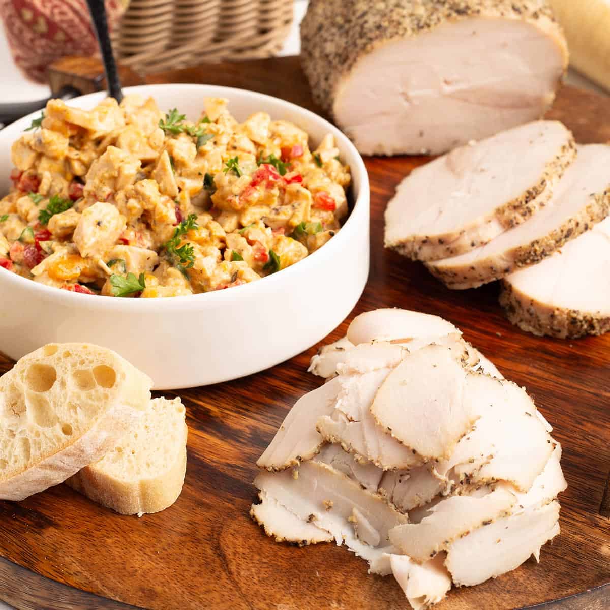 Low roasted turkey breast on a wooden serving board with thick peppered slices, thin shaved slices, a bowl of creamy turkey salad garnished with parsley, and toasted baguette rounds.