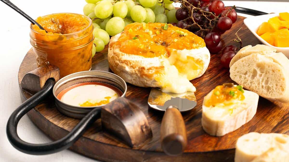 Wooden serving board holding baked brie melting under Mango Jalapeno Jam (No Sugar Added), with an open jar of jam, grapes, crusty bread, and a bowl of diced mango.