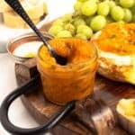 Close-up of a small mason jar of Mango Jalapeno Jam (No Sugar Added) with a black spoon lifting the golden jam, set on a wood board with grapes and baked brie behind it.