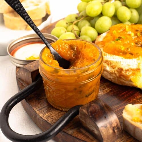Close-up of a small mason jar of Mango Jalapeno Jam (No Sugar Added) with a black spoon lifting the golden jam, set on a wood board with grapes and baked brie behind it.