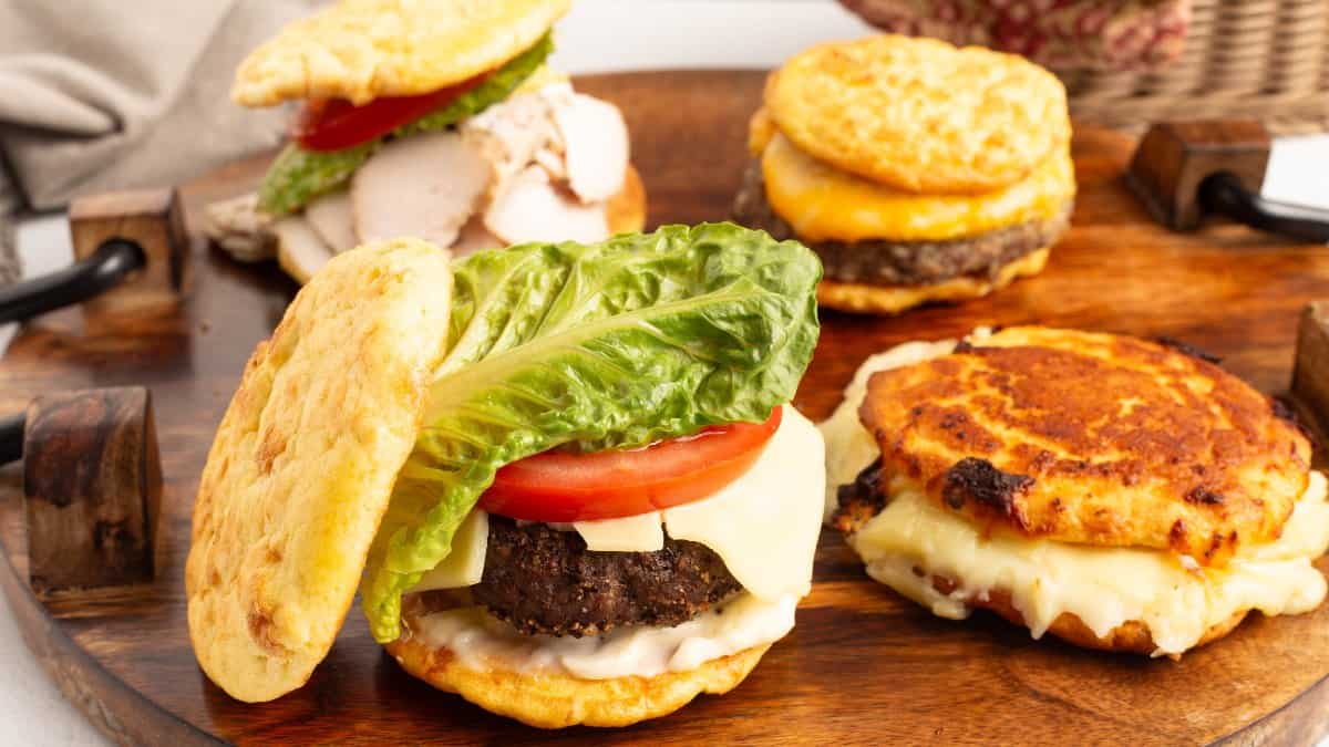 Low Carb Burger Buns holding a beef patty with melted white cheese, a tomato slice, and green leaf lettuce, held in two hands.