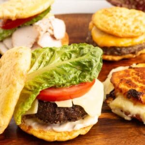Close up of a Low Carb Burger Buns cheeseburger with melted cheese, a thick tomato slice and a romaine lettuce leaf, with more burgers on a wooden board in the background.