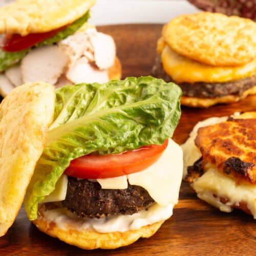 Close up of a Low Carb Burger Buns cheeseburger with melted cheese, a thick tomato slice and a romaine lettuce leaf, with more burgers on a wooden board in the background.
