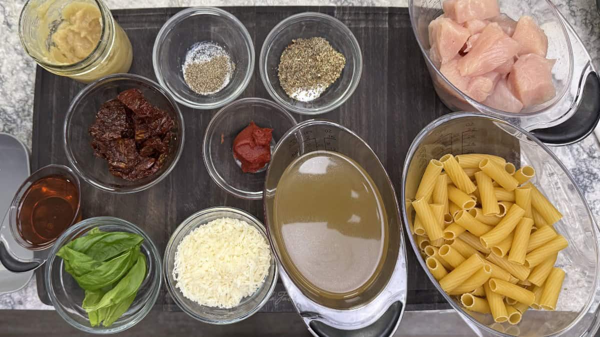 Overhead shot of ingredients for One-Pot Marry Me Chicken (Pressure Cooker), including raw chicken cubes, rigatoni pasta, sun dried tomatoes, chicken broth, cream, shredded parmesan, basil, tomato paste, sherry, salt, pepper, and Italian seasoning in small bowls.