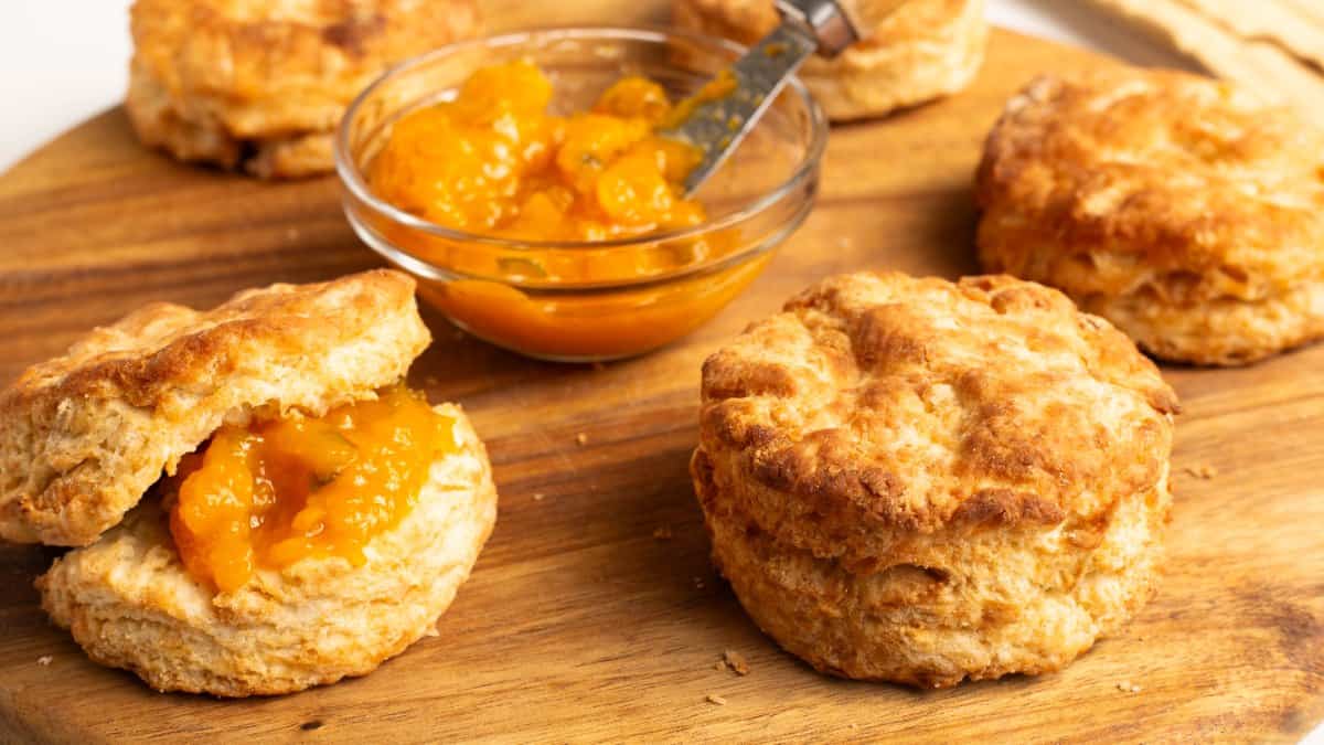 Several golden Air Fryer Homemade Biscuits on a wooden board, one split open and filled with peach jam, with a bowl of jam and a spoon nearby.