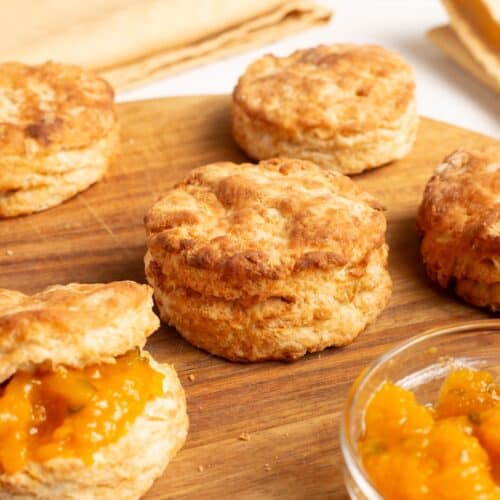 A square crop of Air Fryer Homemade Biscuits on a wooden board, one halved and spread with peach jam beside a small glass jam bowl.