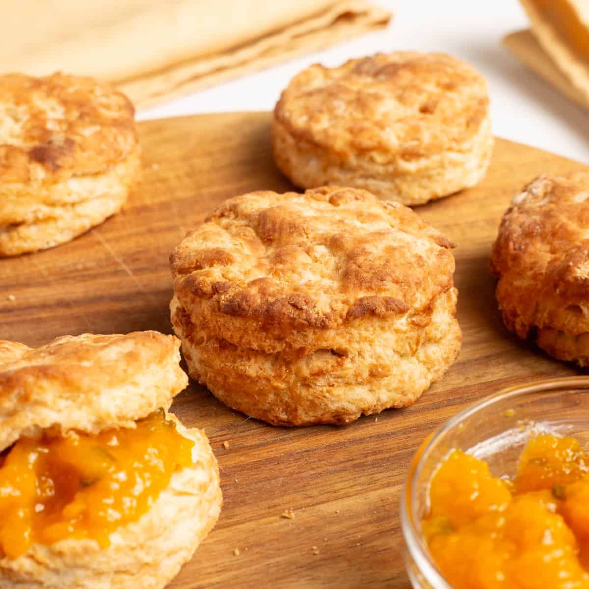A square crop of Air Fryer Homemade Biscuits on a wooden board, one halved and spread with peach jam beside a small glass jam bowl.