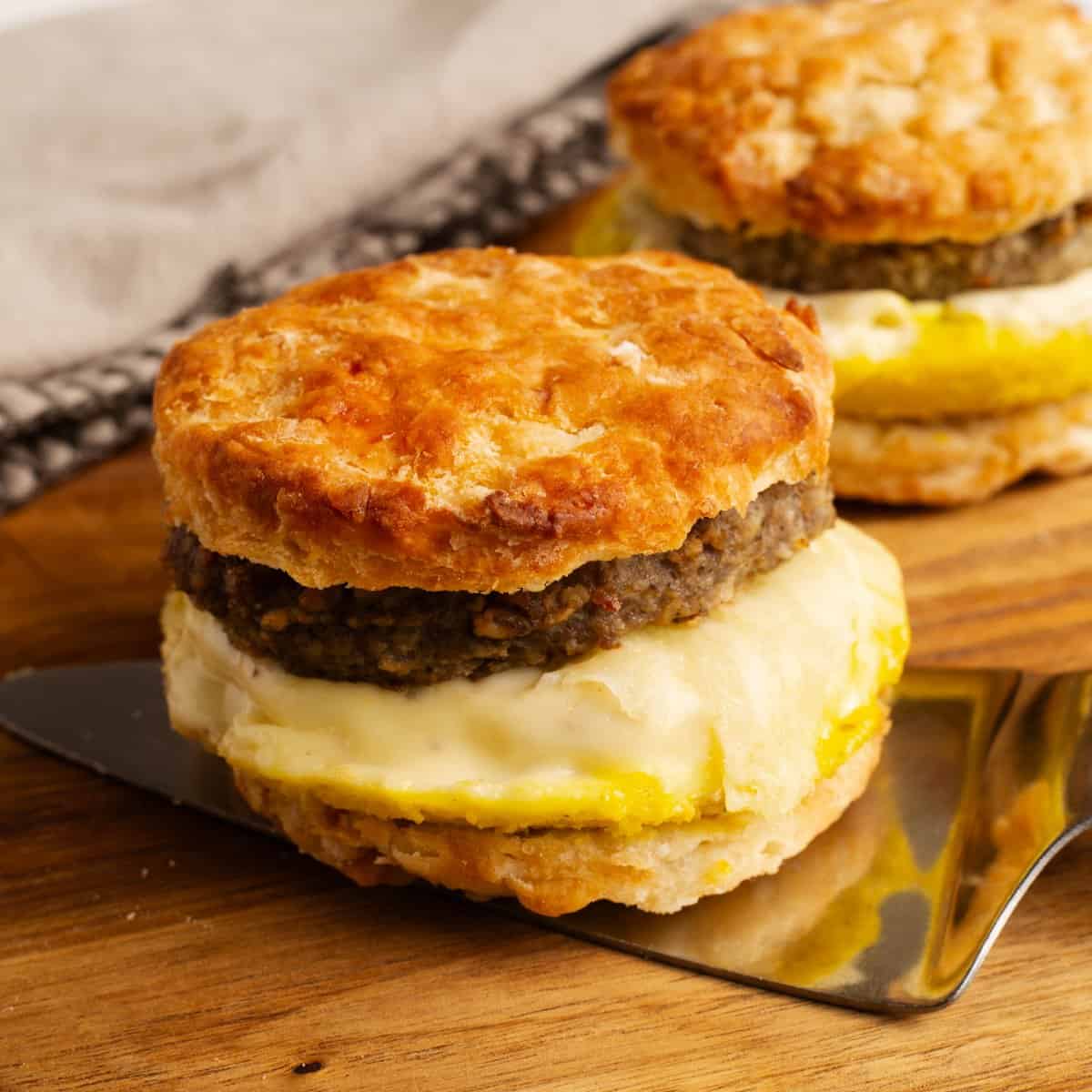 A Breakfast Sausage and Egg Biscuit cut in half, showing a sausage patty and fried egg between two flaky buttermilk biscuit halves, with a second biscuit sandwich in the background.