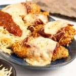 Air Fryer Chicken Parmesan on a blue oval plate, four breaded cutlets topped with melted mozzarella, marinara and herbs, served alongside angel hair pasta with a spoonful of sauce.