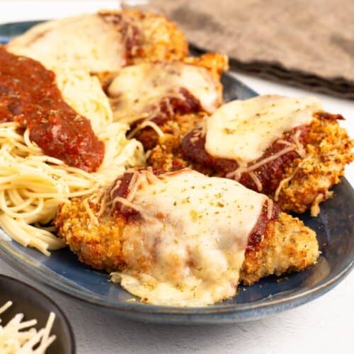 Air Fryer Chicken Parmesan on a blue oval plate, four breaded cutlets topped with melted mozzarella, marinara and herbs, served alongside angel hair pasta with a spoonful of sauce.