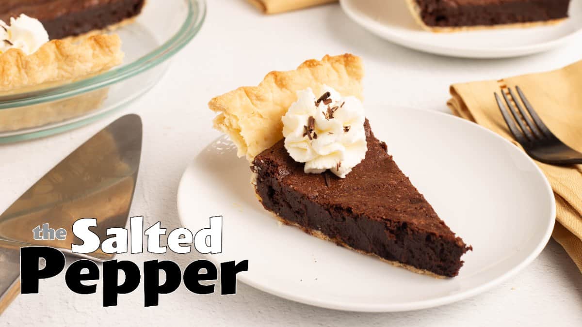 A slice of Mississippi Mud Pie on a white plate, topped with a swirl of Chantilly cream and chocolate shavings, with a piece of the flaky crust edge standing up behind it and the rest of the pie in a glass pie plate in the background.