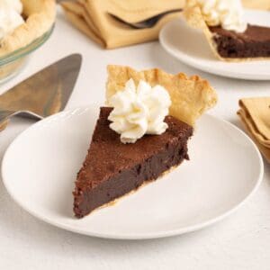A slice of Mississippi Mud Pie on a white plate with a rosette of Chantilly cream and a standing piece of flaky crust, with a pie server and a second slice nearby.