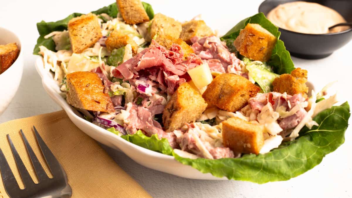 Reuben Salad served in a white oval bowl lined with romaine leaves, filled with creamy slaw, shaved corned beef, cubes of Swiss cheese, and golden rye croutons. A dark bowl of Thousand Island dressing and a fork on a gold napkin sit alongside.