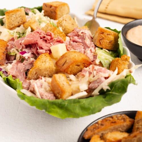 Close view of Reuben Salad in a white bowl lined with romaine, showing shaved corned beef, Swiss cheese cubes, shredded cabbage in creamy dressing, and toasted rye croutons. A bowl of Thousand Island dressing sits behind it and a bowl of extra croutons in front.